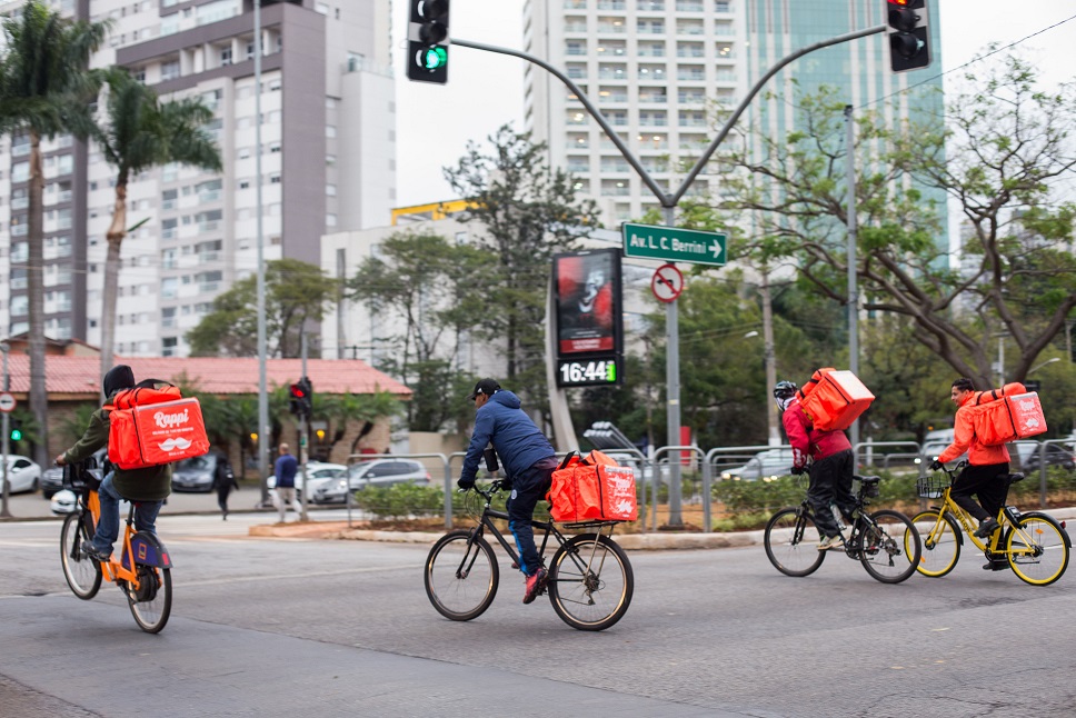 Quatro entregadores em cima de bicicletas em uma rua com bags da Rappi