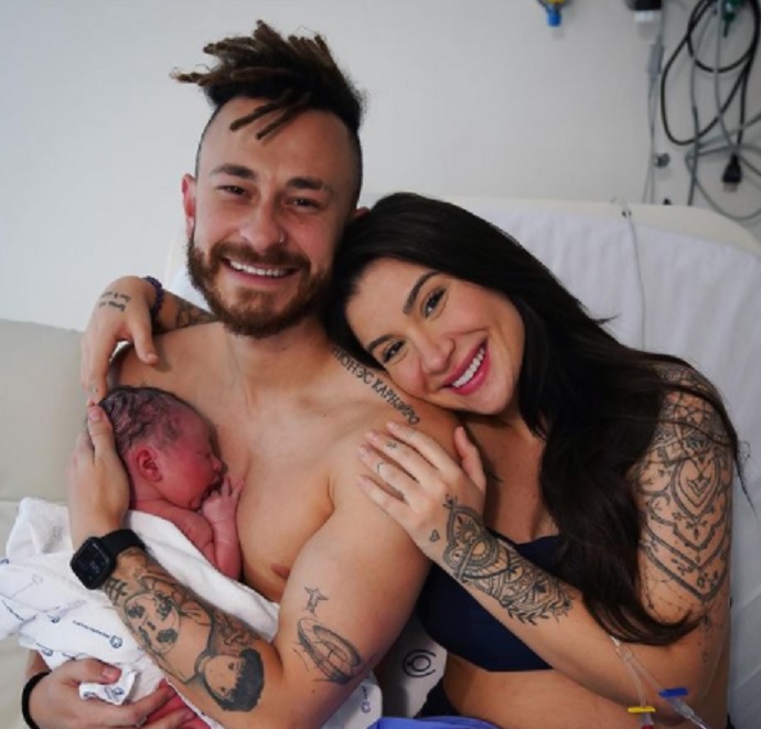 Fred e Boca Rosa com o bebê recém-nascido no colo. Ela está abraçada nele, ele sem camisa e o bebê enrolado em um cobertor branco
