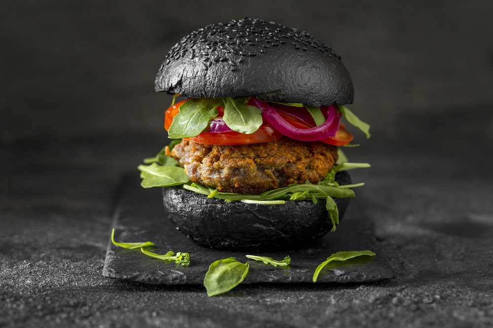 Imagem de frente de um sanduíche de hambúrguer vegetariano com pão preto colocado uma mesa cor de chumbo