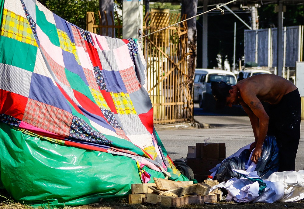Homem em situação de rua se agacha e remexe o lixo ao lado de uma barraca formada por cobertores