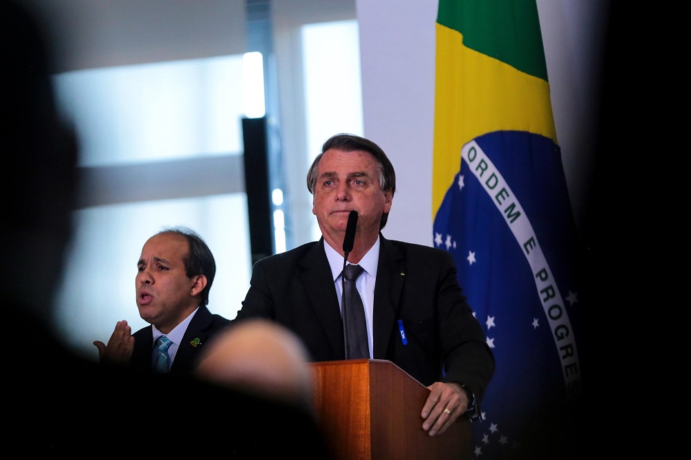De traje social, o presidente Jair Bolsonaro discursa com a bandeira do Brasil atrás e seu intérprete de sinais do lado