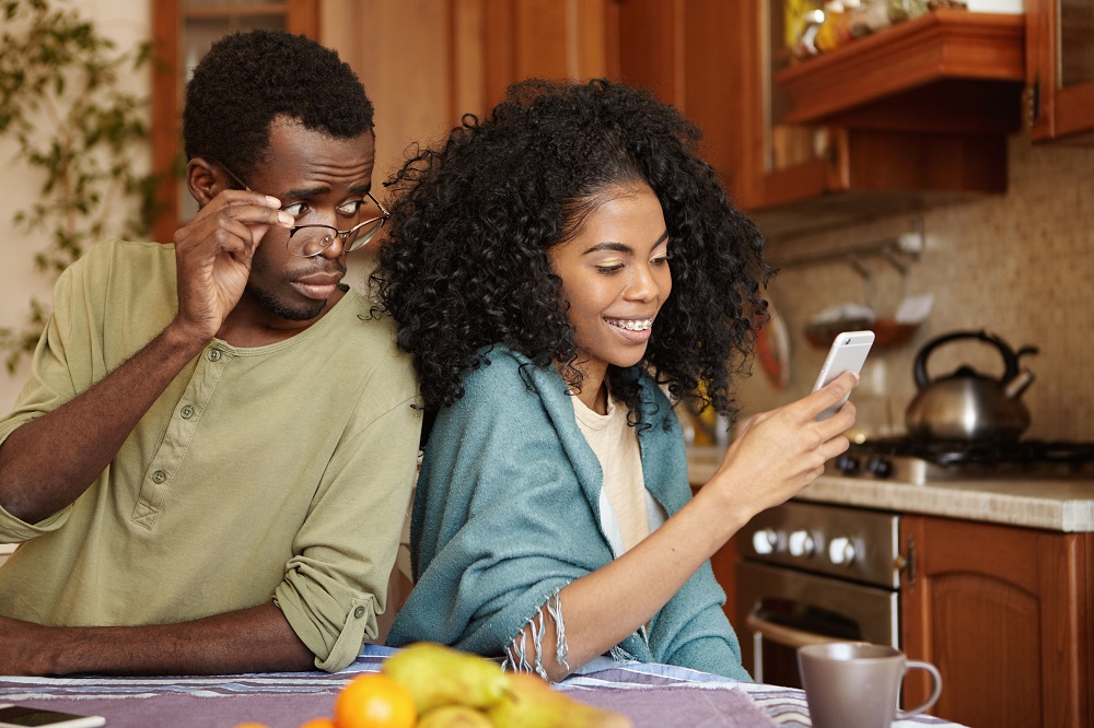 Jovem casal negro lado a lado na cozinha; o homem segura os óculos e tenta ver o que a mulher olha sorridentemente para o celular