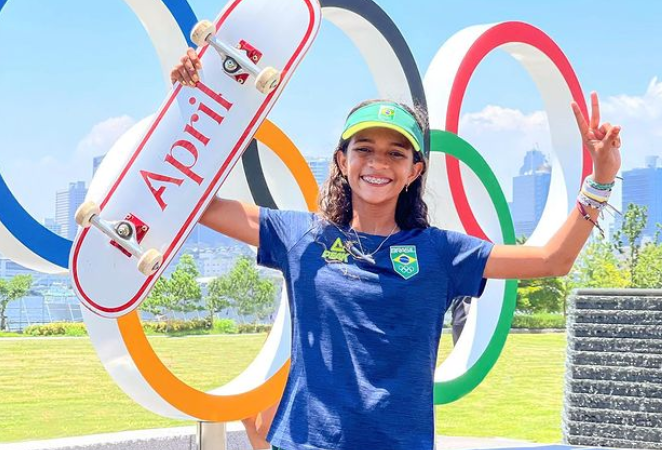 Rayssa Leal está de camisa azul e boné verde, erguendo um skate com o braço com o símbolo olímpico ao fundo