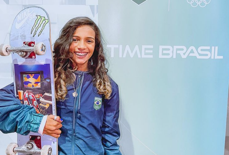 Rayssa Leal tem cabelos castanhos longos e segura um skate colorido e sorri ao fundo de uma parede com os escritos 'Time Brasil'