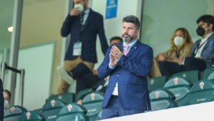 Homem de terno azul em pé em um estádio de futebol com cadeiras. Tem as mãos juntas e olha para o horizonte. Algumas pessoas estão atrás sentadas e em pé