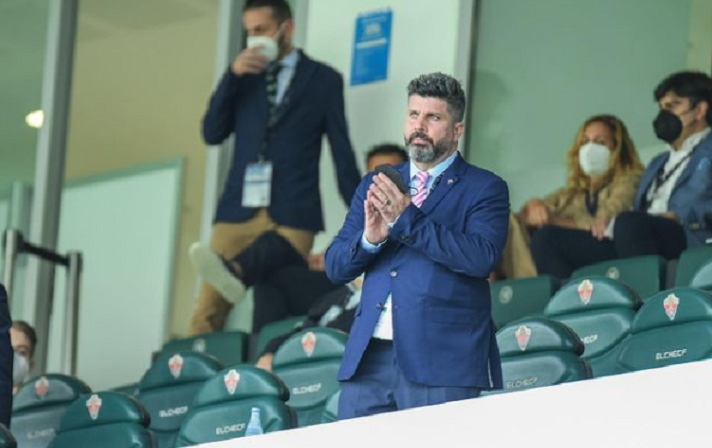 Homem de terno azul em pé em um estádio de futebol com cadeiras. Tem as mãos juntas e olha para o horizonte. Algumas pessoas estão atrás sentadas e em pé