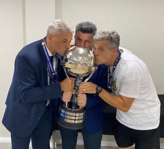 Tres hombres besando una copa de una liga de fútbol.