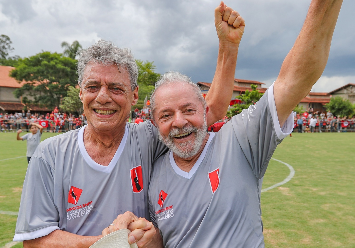 Dois homens com camisetas cinzas de futebol abraçados com as mãos para cima com um campo de futebol atrás