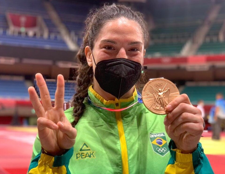 Com o uniforme do Brasil, o cabelo preso com rabo de cavalo, Mayra Aguiar segura a medalha de bronze com a mão esquerda e faz sinal de três com a mão direita