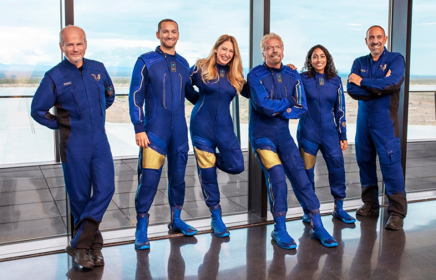 Seis pessoas em pé posando para uma foto com uniformes azuis da companhia Virgin Galactic. Eles viajarão ao espaço