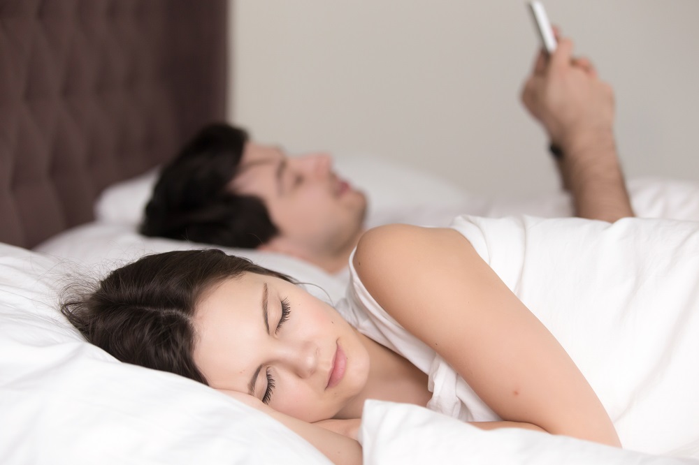 Em uma cama de casal, jovem mulher dorme enquanto seu companheiro mexe no celular; ambos são brancos