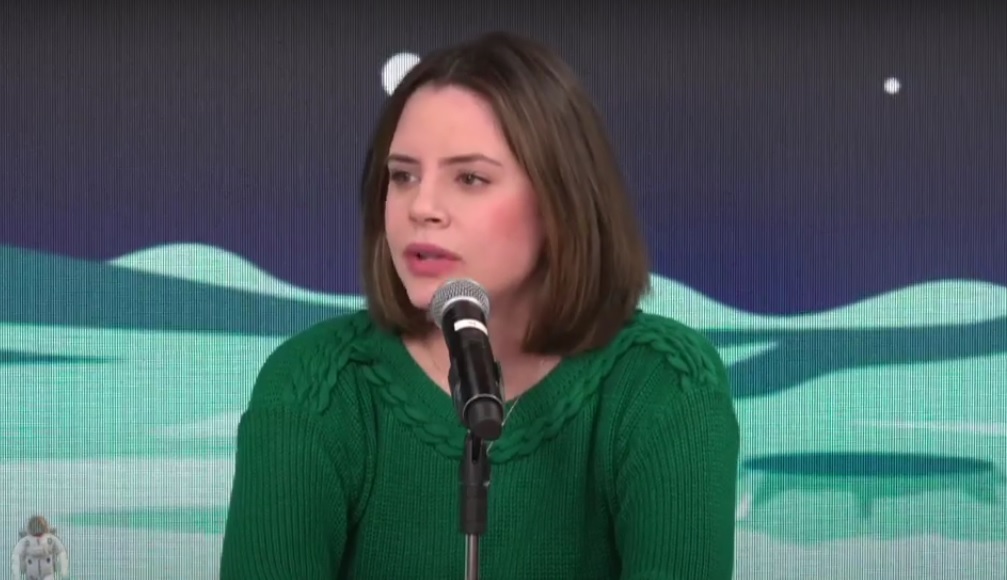 Zoe Martinez, mulher branca de cabelos curtos castanhos e blusão verde de lã, fala ao microfone