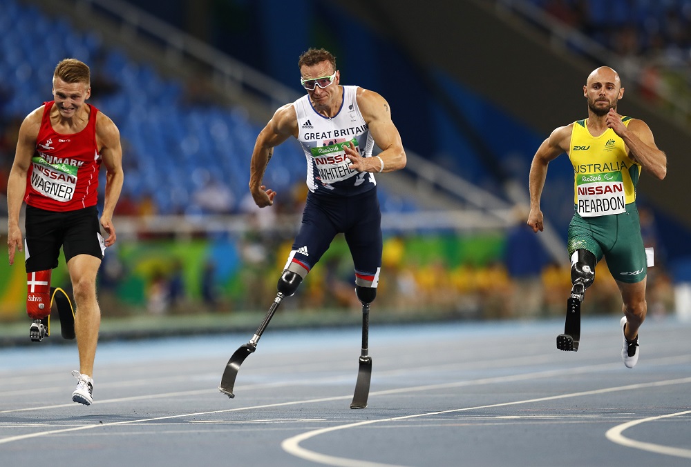 Três atletas amputados disputam a prova dos 100 m: um britânico, de branco, que lidera em uma pista central, um dinamarquês de vermelho, à esquerda, e um australiano, de amarelo, à direita