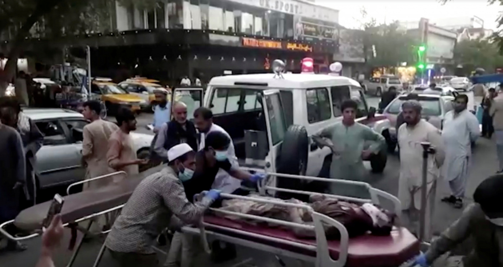Pessoas amontoadas em cima de uma maca com alguém deitado e uma ambulância