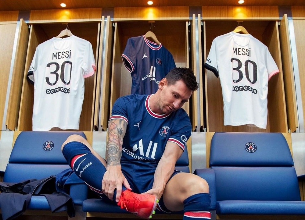 Sentado em um banco do vestiário do PSG, vestido com o uniforme todo azul do clube, Messi flexiona a perna direita para calçar a chuteira laranja; três camisas estão penduradas atrás dele: duas brancas, com o número 30 e o nome de Messi nas costas e uma azul