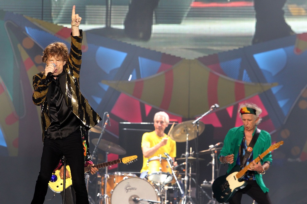 À frente do palco, Mick Jagger canta segurando o microfone com a mão direita e ergue o braço esquerdo, enquanto, atrás dele, o guitarrista Keith Richards e o baterista Charlie Watts tocam seus instrumentos