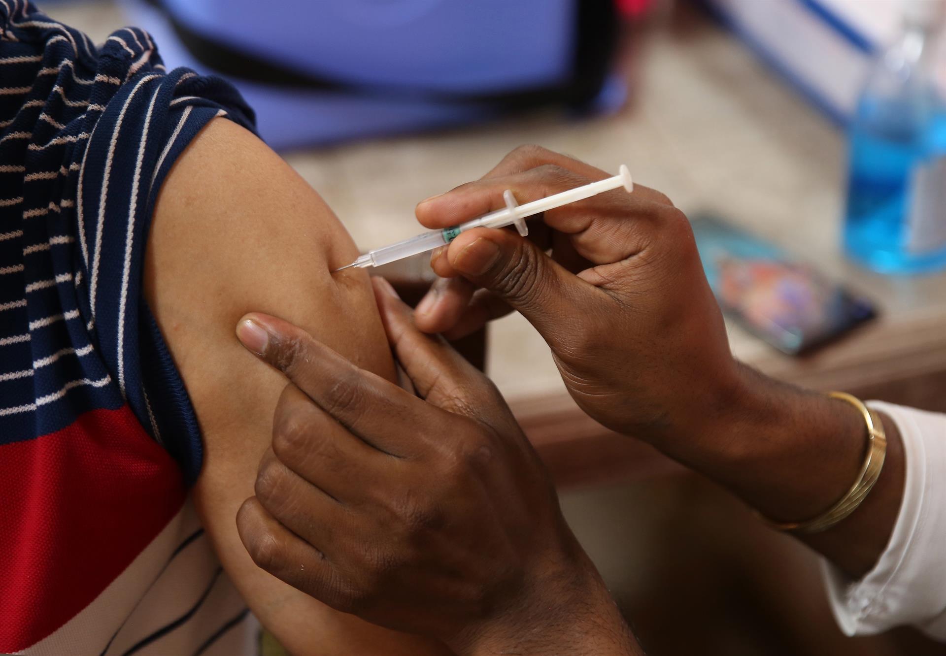 Pessoa colocando agulha no braço de outra, vacinando contra a Covid-19. Na foto só mostra a mão e o braço das duas.