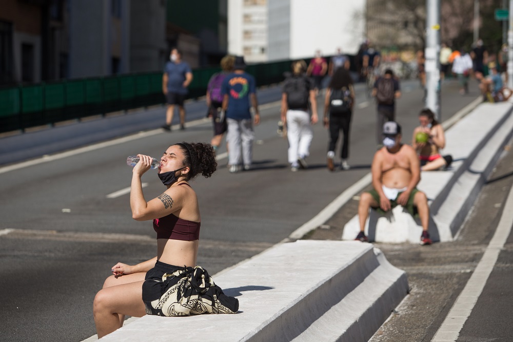 Mulher de top e saia, sentada no canteiro central do Minhocão, bebe água; há um homem de boné e sem camisa a alguns metros dela, e um grupo de pessoas andando de costas para ela em uma das vias