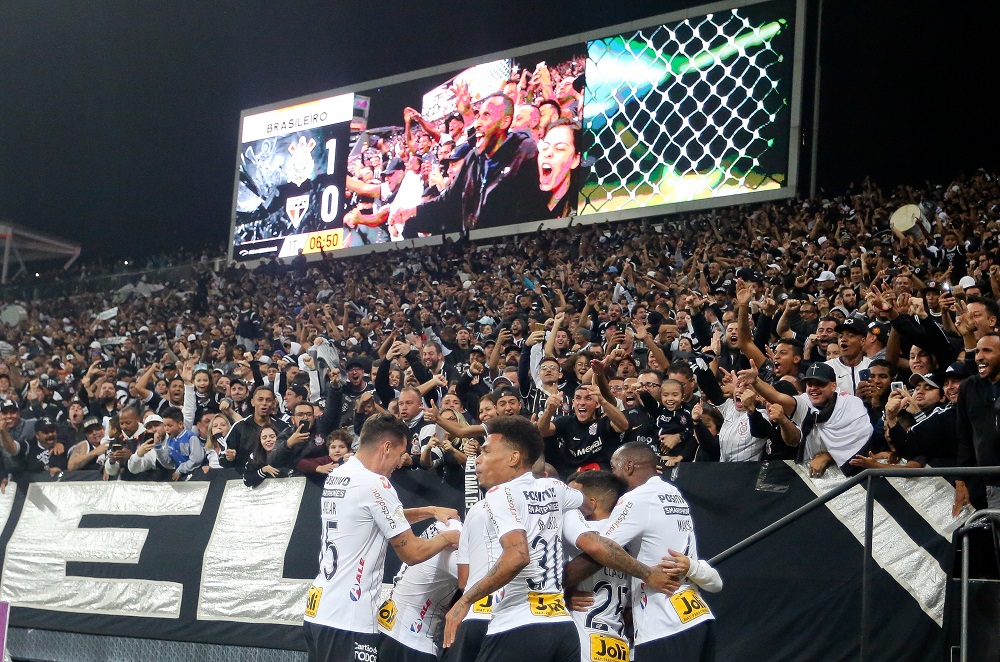 Seis jogadores do Corinthians festejam um gol perto da arquibancada, torcedores fazem festa e placar mostra Corinthians 1 x 0 São Paulo