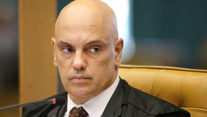 Ministro Alexandre de Moraes olhando com os olhos de canto, sentado em cadeira amarela, com terno vermelho, camisa branca e gravata bordô com bolinhas