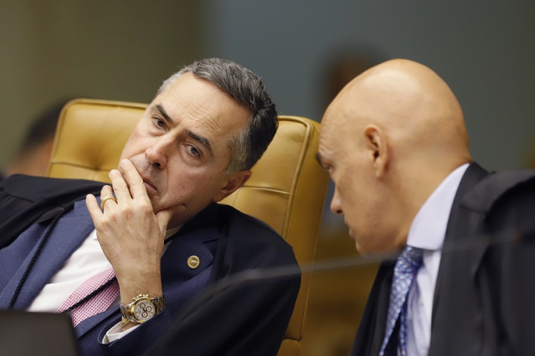Luís Roberto Barroso estica o pescoço para ouvir Alexandre de Moraes, que está à sua esquerda (ambos estão de toga e em trajes sociais)