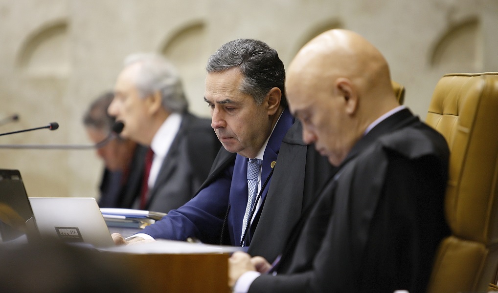 O ministro do STF Alexandre de Moraes olha para o seu celular, enquanto o colega Luís Roberto Barroso se concentra em seu notebook; ambos estão sentados em uma bancada, e próximo a eles, desfocado, aparece Ricardo Lewandowski