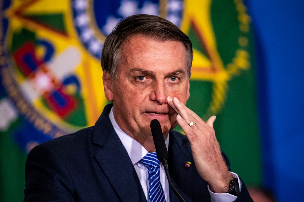 Jair Bolsonaro coçando ao lado do nariz, atrás de um microfone e na frente de uma bandeira do Brasil. Usa terno e gravata.