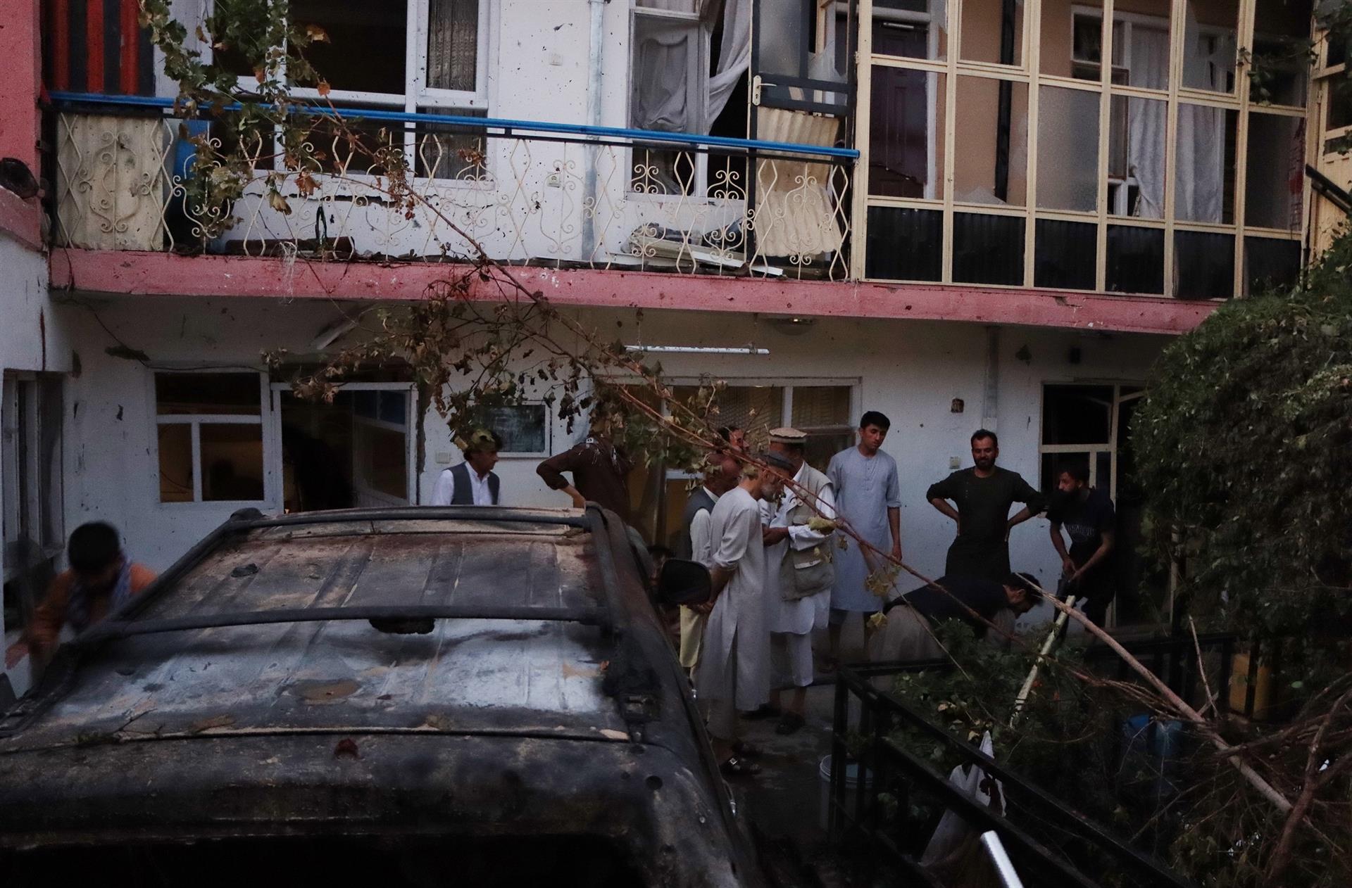 Pessoas em volta de um carro, árvores e outras estruturas destruídas por bomba no Afeganistão. Atrás, janelas de uma casa