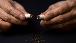 As mãos de um homem seguram cada uma a metada de um cigarro partido ao meio, com um pouco de tabaco sobre uma mesa escura