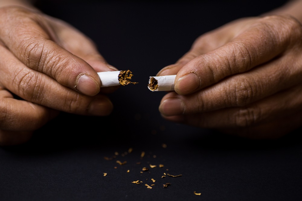 As mãos de um homem seguram cada uma a metada de um cigarro partido ao meio, com um pouco de tabaco sobre uma mesa escura