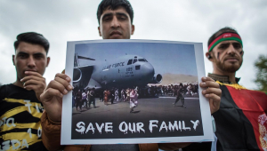 Três afegãos protestam em Paris, sendo que um deles (o do meio) segura com as duas mãos um cartaz com um avião e a frase "Salvem nossas famílias"