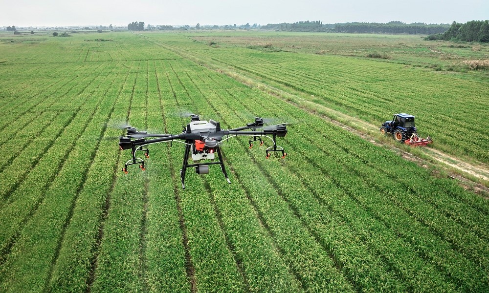 Imagem aérea mostra um drone sobrevoando um extenso campo agrícola, por onde passa um trator
