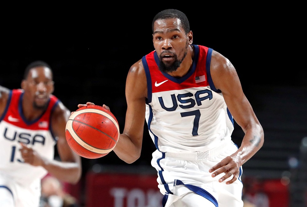 Com uniforme branco da seleção americana de basquete, o jogador Kevin Durant (de bigode e cavanhaque) maneja a bola antes de batê-la no chão