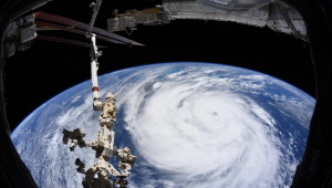 Um furacão visto do espaço. Nuvens brancas em formato de furacão e azul em volta, no planeta Terra. Um satélite também aparece na foto