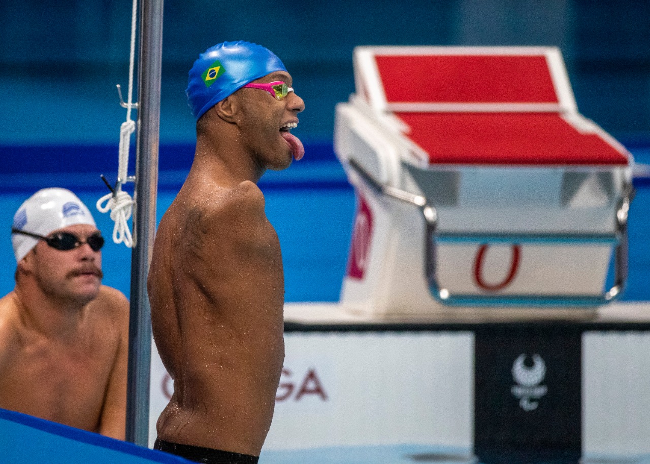 Gabriel, nadador paralímpico, com a língua para fora, touca de natação azul e óculos de natação vermelhos