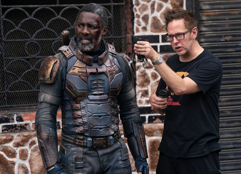 O diretor James Gunn, de camiseta preta e bermuda, orienta o ator Idris Elba, trajado como o vilão Bloodsport