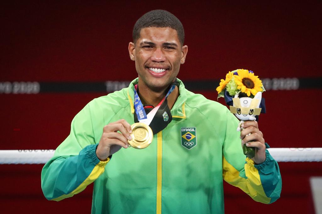 Com o uniforme do Time Brasil, Hebert Conceição dá largo sorriso,exibe a medalha de ouro com a mão direita e segura o mascote olímpico e um ramo de flores com a esquerda
