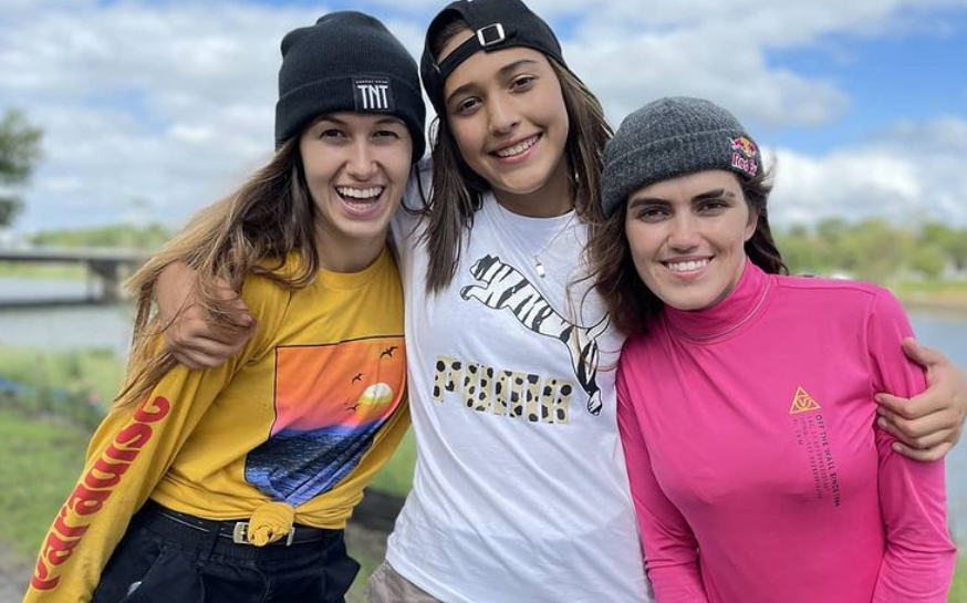 Dora Varella, Isa Pacheco e Yndiara Asp aparecem abraçadas sorrindo com uma paisagem natural verde e azul ao fundo