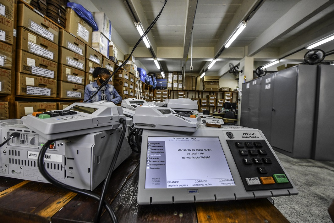 Duas urnas eletrônicas em evidência dentro de um espaço com várias caixas e outras urnas atrás