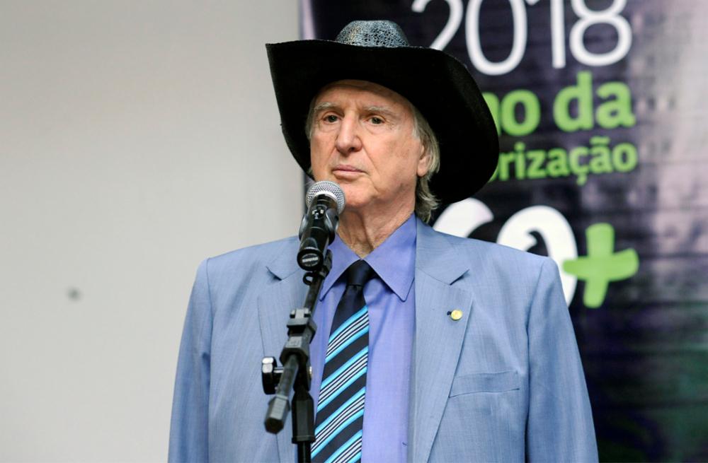 Sérgio Reis de terno azul e chapéu preto em frente a um microfone