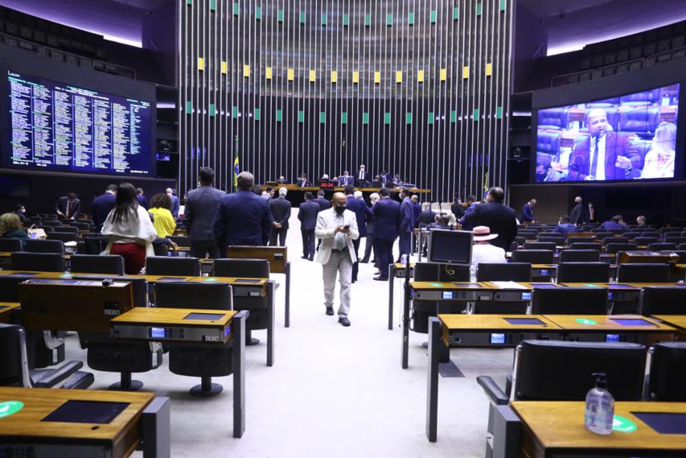 Imagem do plenário da Câmara vista do fundo, com muitos deputados de costa, um no telão e um saindo