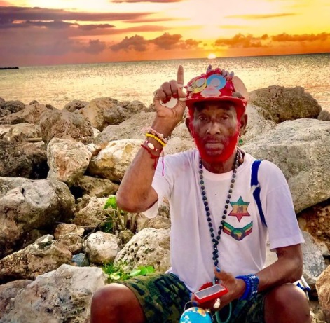 Homem idoso sentado em pedras em uma praia com um boné vermelho, barba pintada de vermelho, camiseta branca e pulseiras e colares coloridos. Ele está com o braço e o dedo indicador direitos para cima.