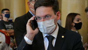 Ministro João Roma falando ao celular e usando máscara de proteção individual. Tem cabelos curtos castanhos e usa terno e óculos de grau