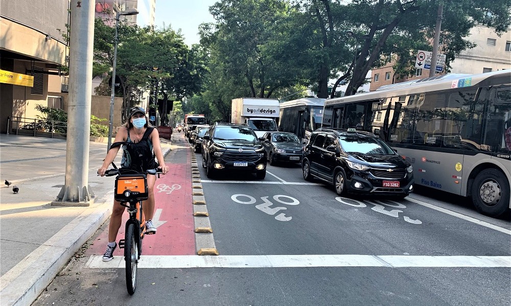 Ao lado de uma pista repleta de carros e ônibus, mulher para sua bicicleta na ciclovia construída ao lado da via para automotores