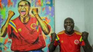 Ao lado de quadro que retrata seu gol, Rincón sorri e vibra com a camisa vermelha da seleção colombiana