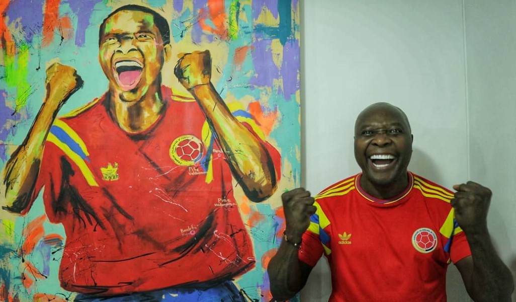 Ao lado de quadro que retrata seu gol, Rincón sorri e vibra com a camisa vermelha da seleção colombiana