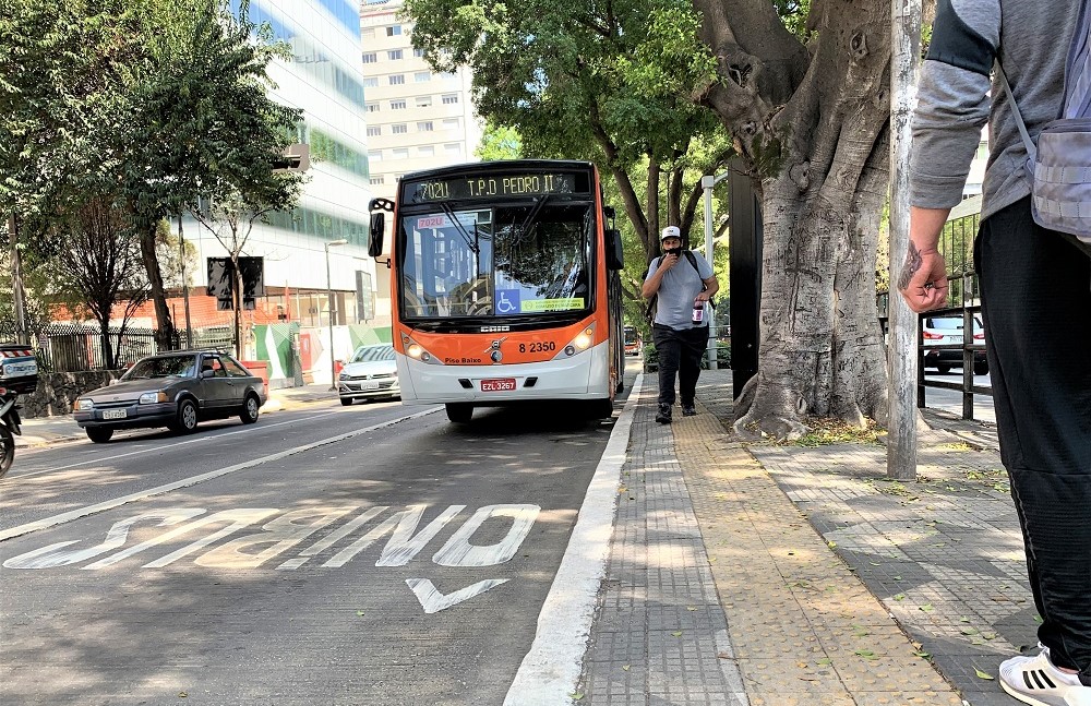 Ônibus laranja, com a localização Pq. Dom Pedro II no letreiro, para no ponto de ônibus, pouco antes da palavra ônibus escrita para indicar o corredor; a via é arborizada, dois carros andam à direita do coletivo e um homem, de boné, mascara, camiseta e calça, anda pela calçada e espaço destinado aos ônibus é limitado