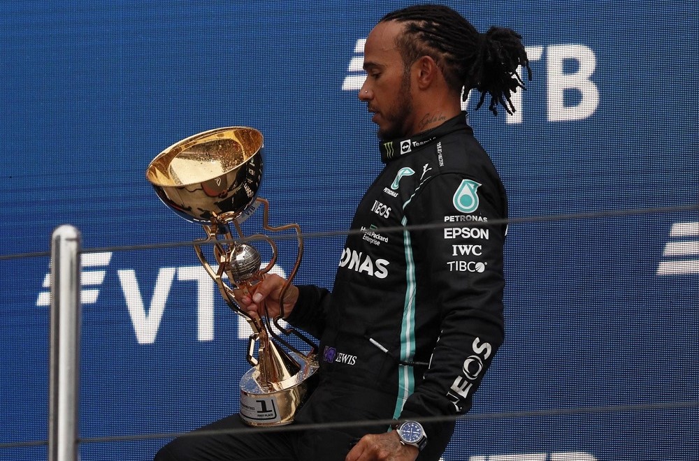 Com o joelho flexionado, vestindo o macacão preto da Mercedes, Lewis Hamilton segura o troféu do GP da Rússia com a mão esquerda