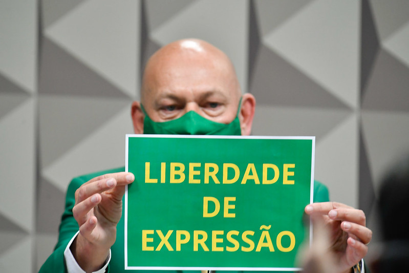 Luciano Hang em sessão na CPI segurando uma placa verde escrito em amarelo 'Liberdade de Expressão'. Usa um terno e uma máscara verdes