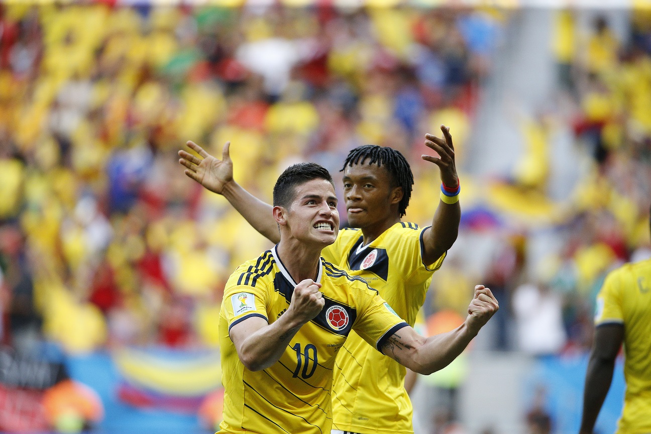 James Rodriguez e colega de equipe comemoram um gol, os dois com os braços ao alto. Usam camisetas amarelas e estão em um estádio